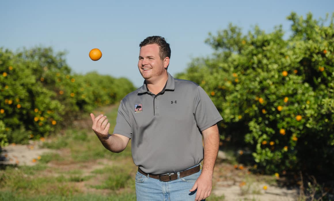 man standing in an orange grove tossing an orange in the air