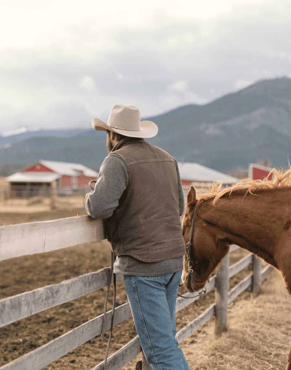 Person in a cowboy hat standing next to a horse at a ranch fence with mountains in the background, offering professional services.