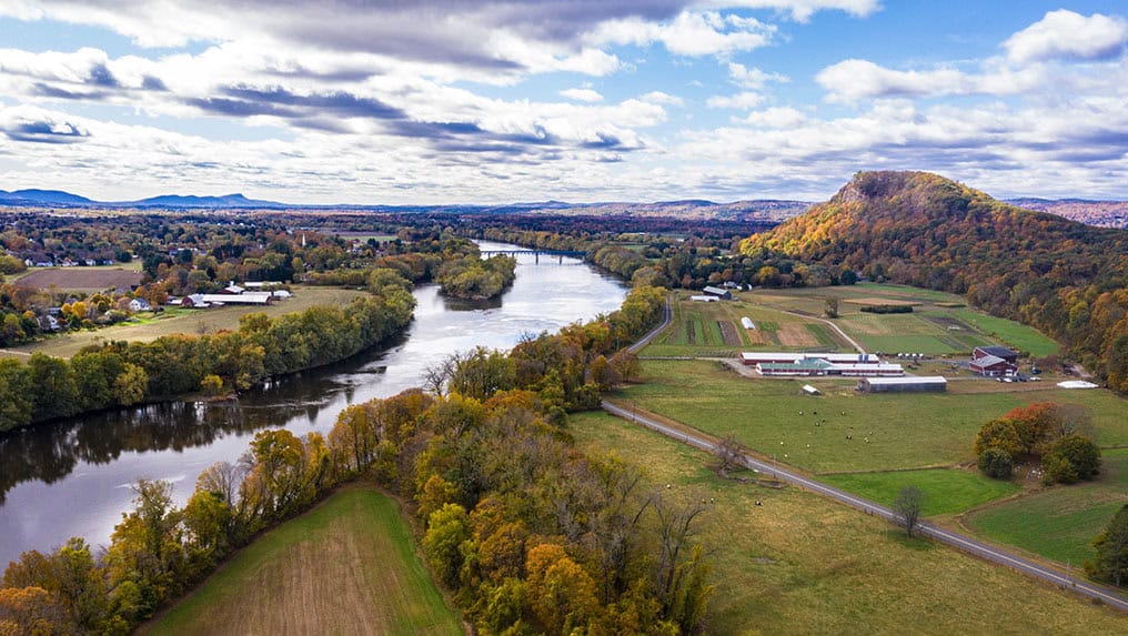 Aerial view of a river meandering through a rural landscape with fields, scattered buildings, and a prominent hill under a partly cloudy sky, highlighting the heartland that could be pivotal in shaping the 2024 Election.