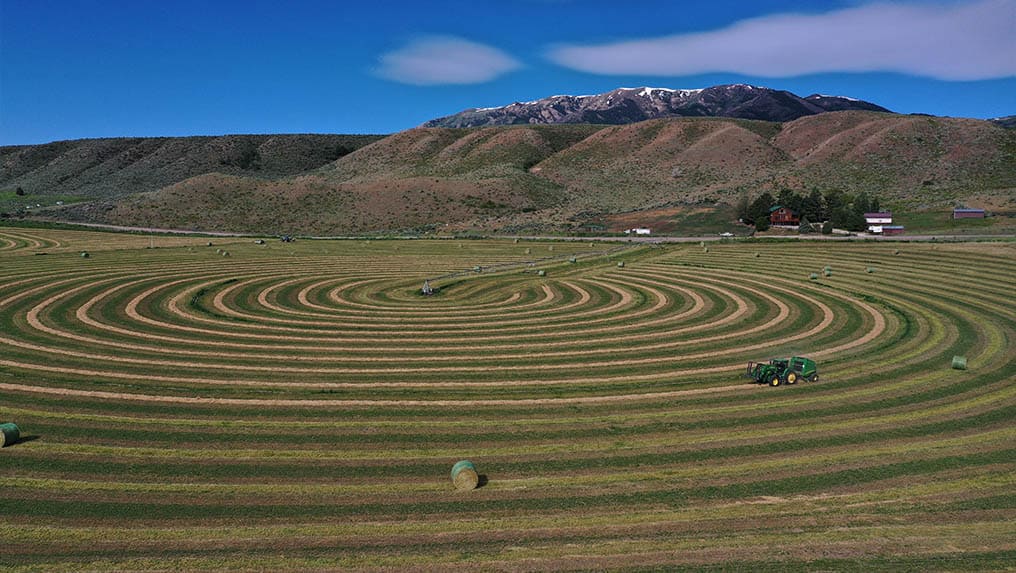 Aerial view of a tractor creating concentric circles in a hay field at the base of rolling hills with mountains in the background, showcasing land as collateral for its agricultural potential.