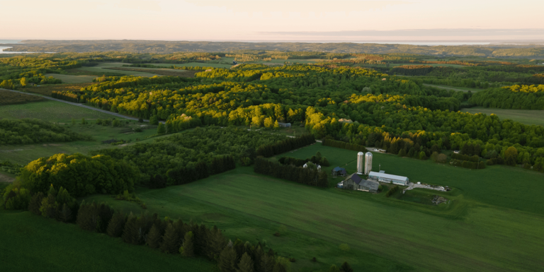 Aerial view of a rural landscape with sprawling green fields, patches of forest, and a farm with silos and buildings under a clear sky, showcasing the vast agricultural land.