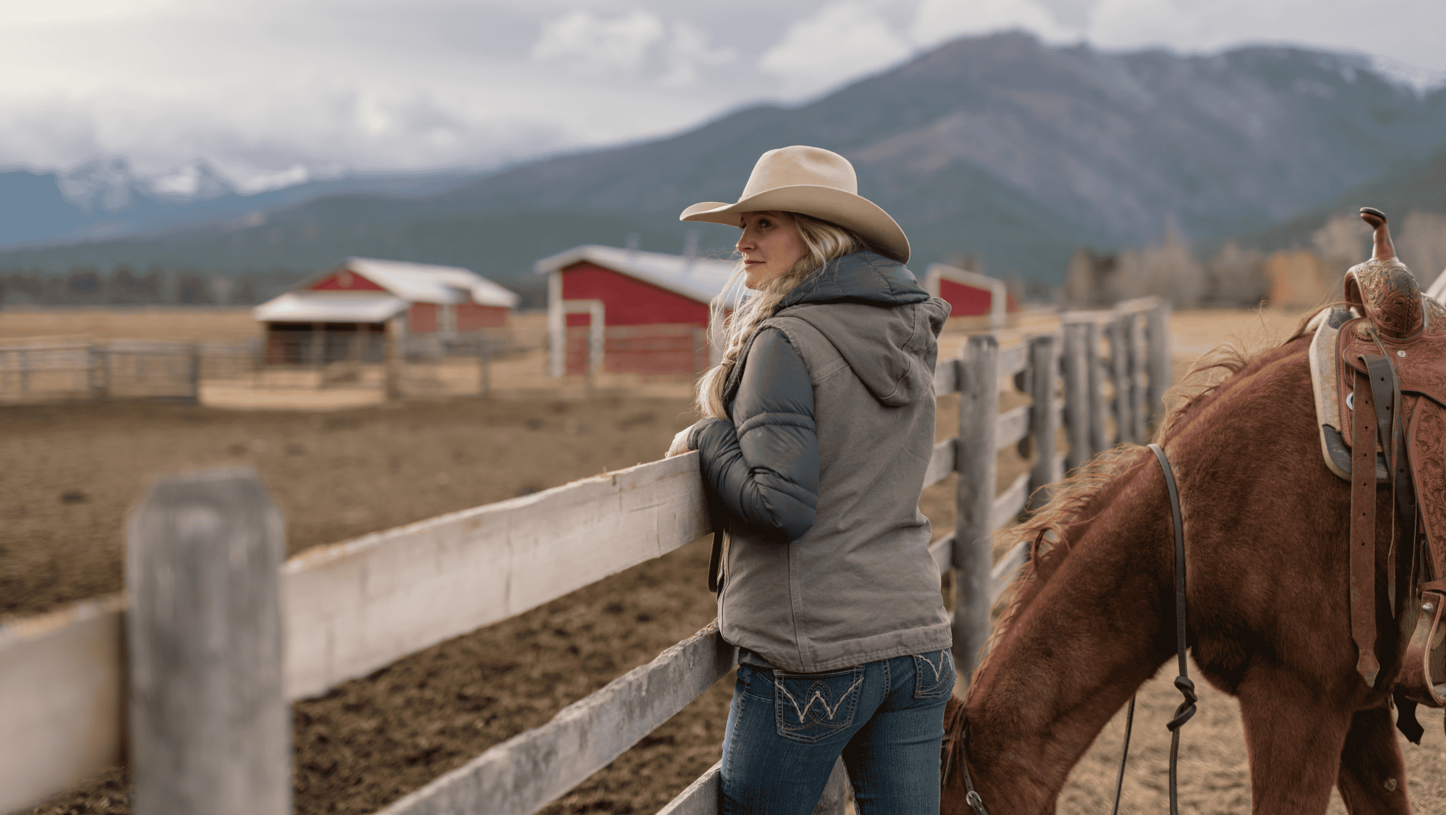 A person in a cowboy hat and jacket stands by a fence next to a saddled horse, with red barns and mountains in the background, embodying the spirit of farmers who benefit from ag loans.