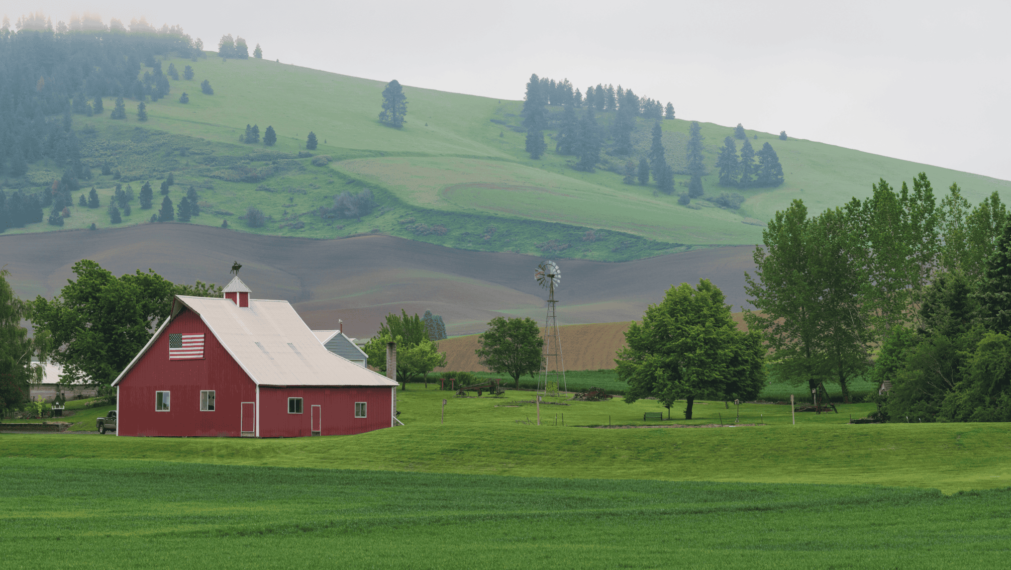 A red barn with an American flag and a windmill stands amidst green fields and rolling hills, symbolizing America's heartland on an overcast day—a scene reflecting the importance of Agricultural Policy in the 2024 Election.