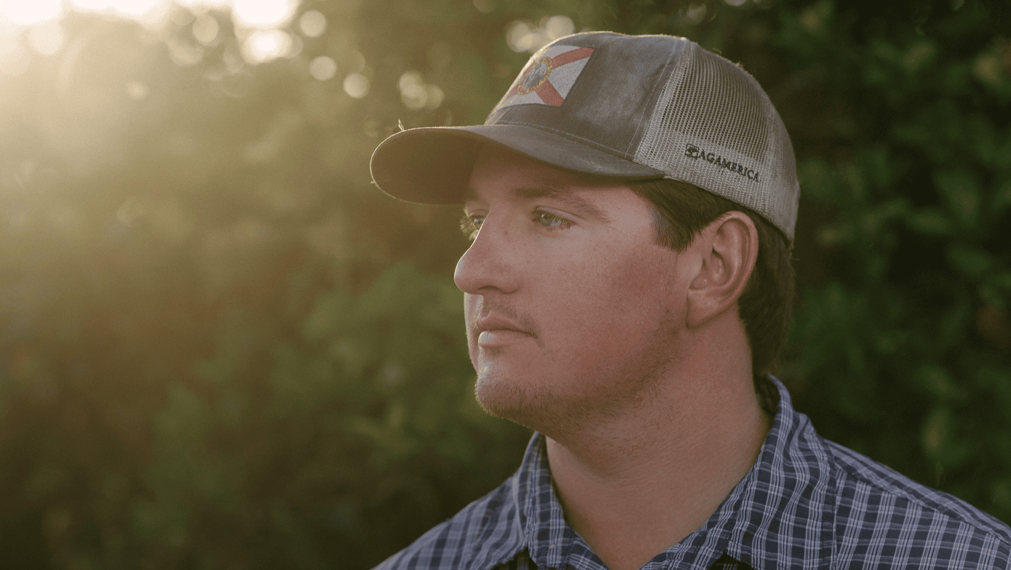 A man with a serious expression, reminiscent of many hardworking U.S. farmers, stands outdoors in a cap and a plaid shirt, against a blurred background of greenery and sunlight.