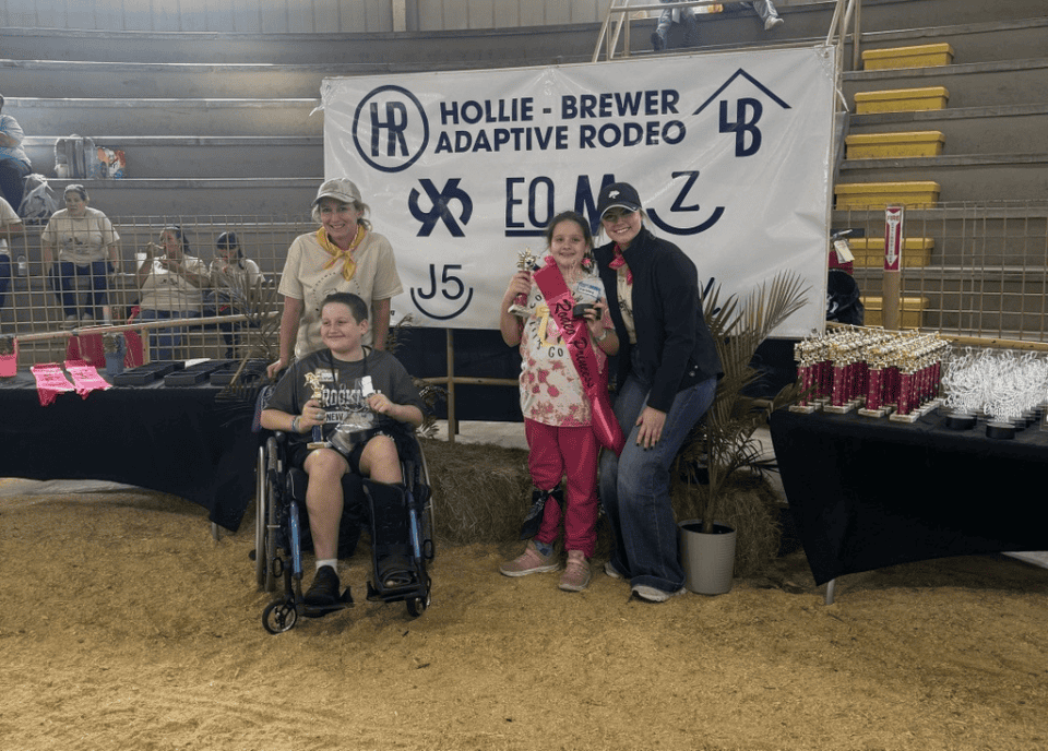 Four people, including Erin Smith, are smiling in front of a banner for the Hollie-Brewer Adaptive Rodeo. One person is in a wheelchair, embodying the spirit of inclusion. Nearby tables showcase trophies shining brightly under the AgAmerican Spotlight.