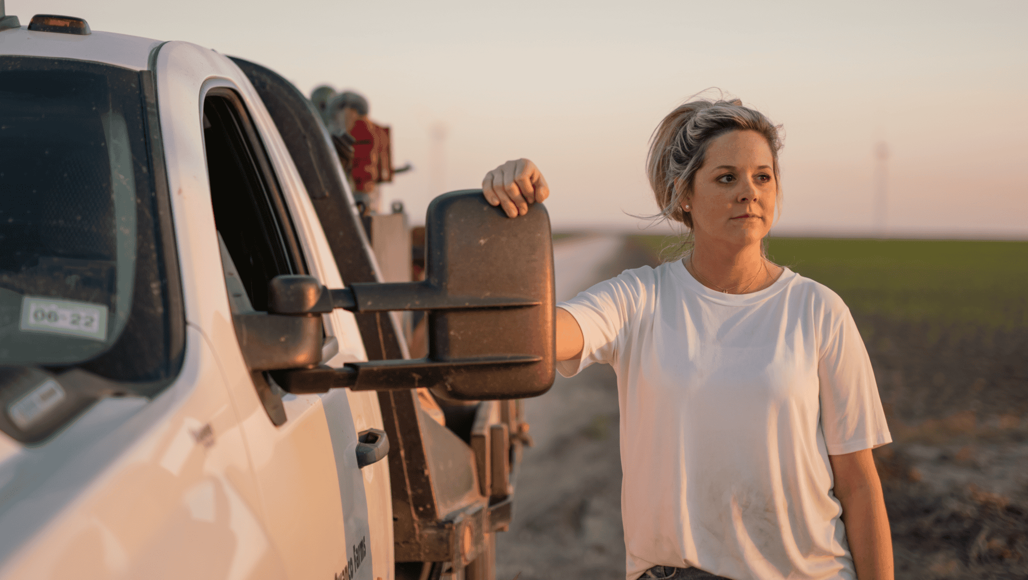 A person in a white shirt stands beside a white truck at sunset in a rural area, holding the side mirror. Fields and roads stretch out behind them, as they ponder the challenges of managing farm debt in this high-rate environment.