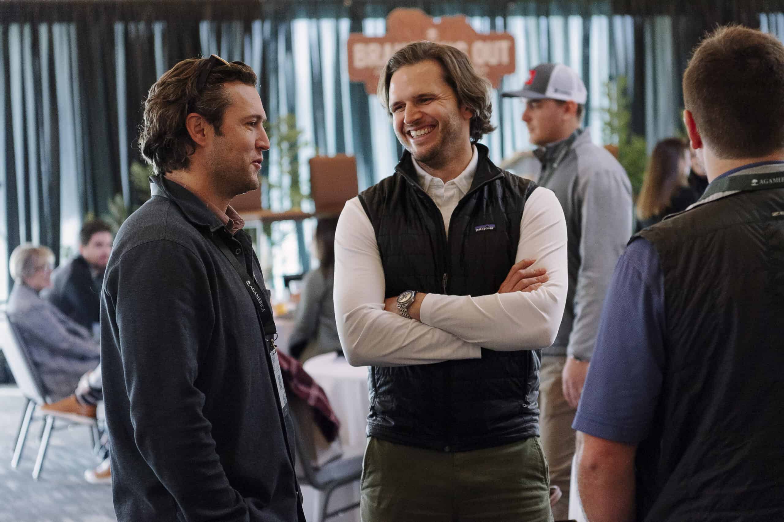 Max Davis and another man, both in casual attire, are engaged in conversation at a social event. Surrounded by tables and mingling guests, they contribute to the relaxed atmosphere with ease and camaraderie.