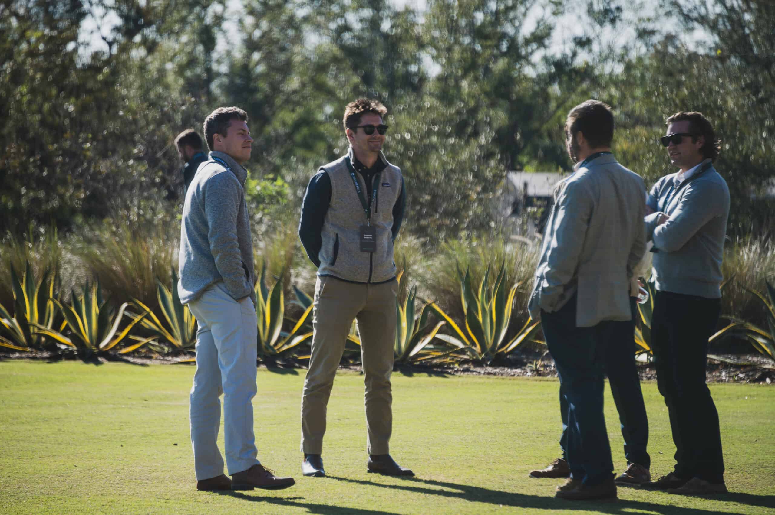 Four men, including Max Davis, stand outdoors on grass, casually conversing amidst a serene setting of plants and trees. Their discussion seems to capture the essence of an AgAmerican Spotlight moment.