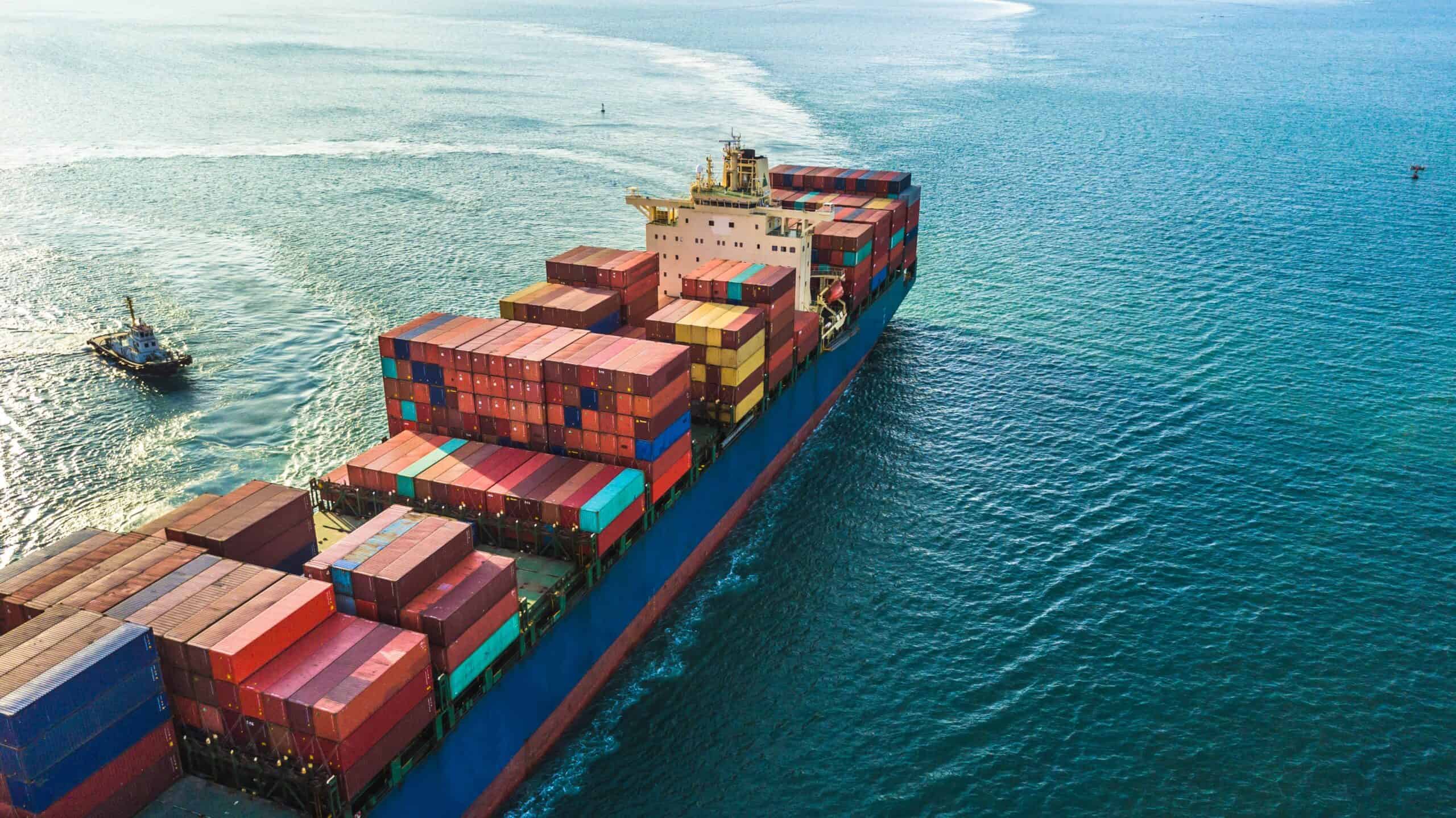 A large cargo ship loaded with colorful containers, crucial for agricultural trade, sails through open blue waters. A smaller boat is visible nearby, navigating the tides of changing tariffs as we approach the 2025 outlook.