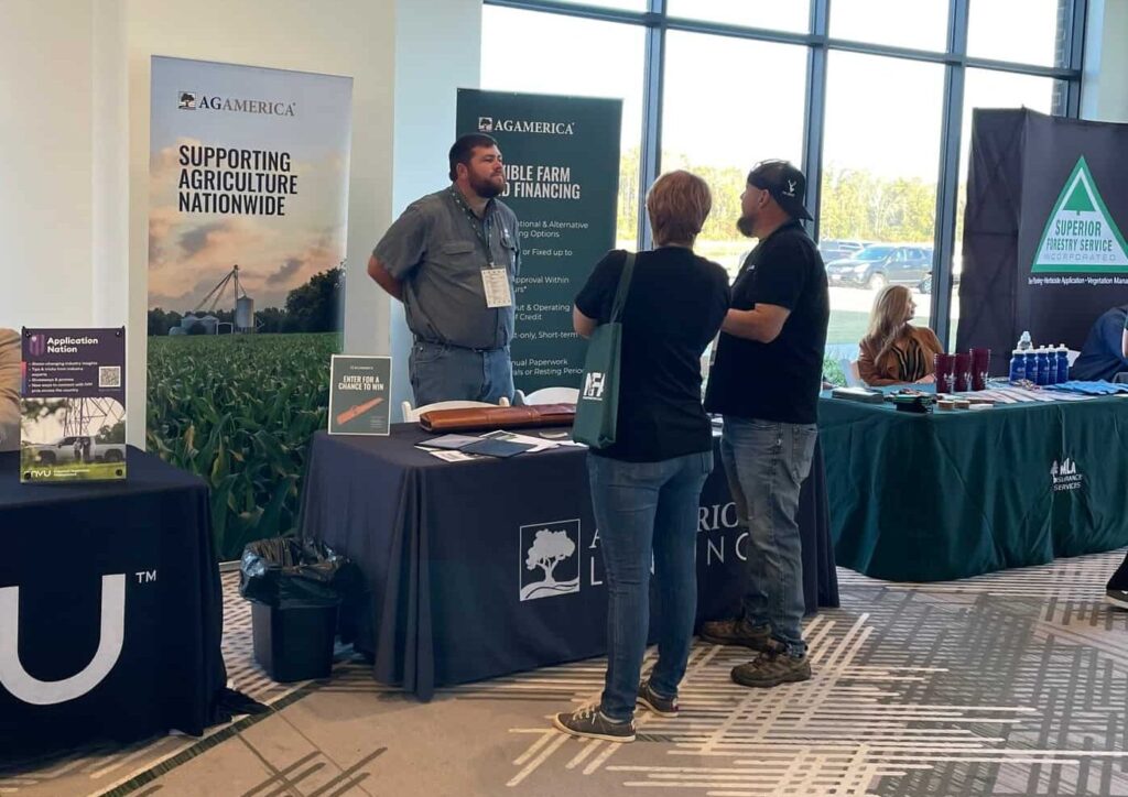 People stand and converse at an indoor event with vendor tables displaying banners about agriculture and finance, including AgAmerica. Large windows provide natural light in the background as Clay Tucker mingles with attendees.