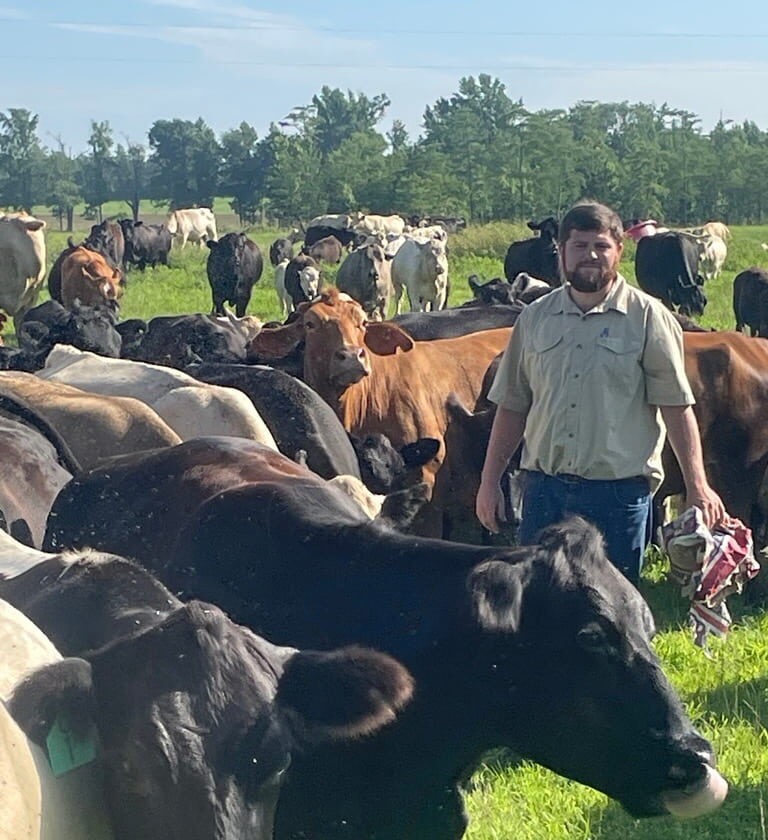 A man stands in a grassy field surrounded by a large herd of cows under a clear blue sky, embodying the spirit of American agriculture championed by AgAmerica.