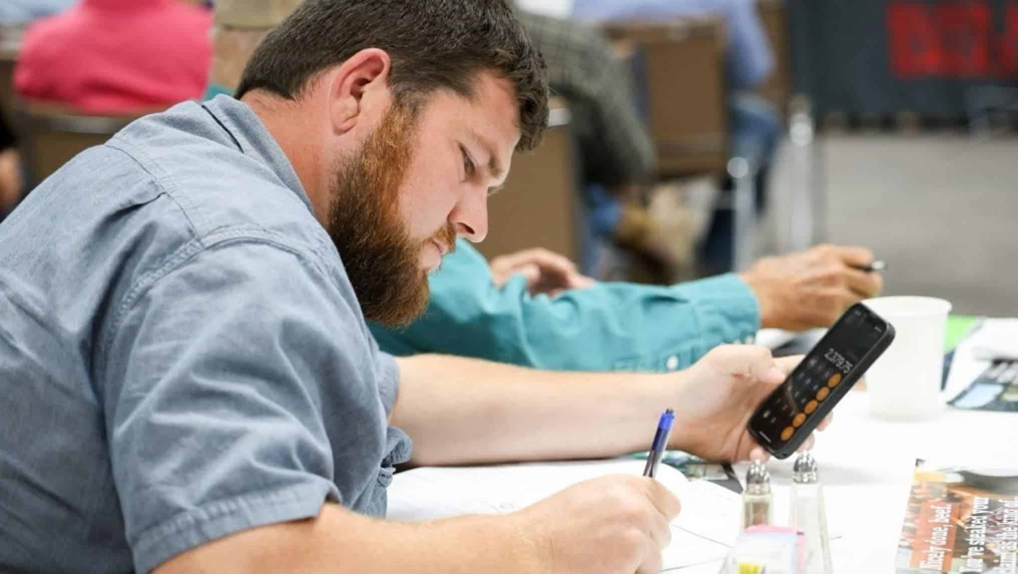 A man sits at a table using a calculator and writing on paper, appearing focused, with AgAmerica documents and other people visible in the background.