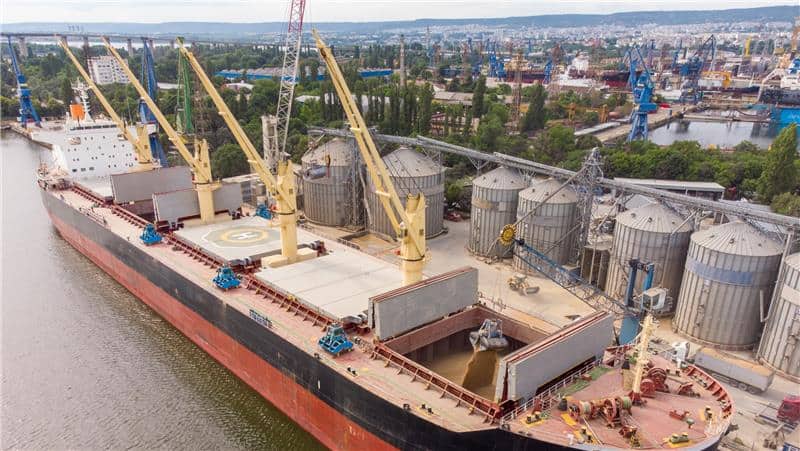 A cargo ship docked at a port is being loaded with grain from silos using cranes and conveyor belts, illustrating the flow of farm trade, with an industrial area and city visible in the background.