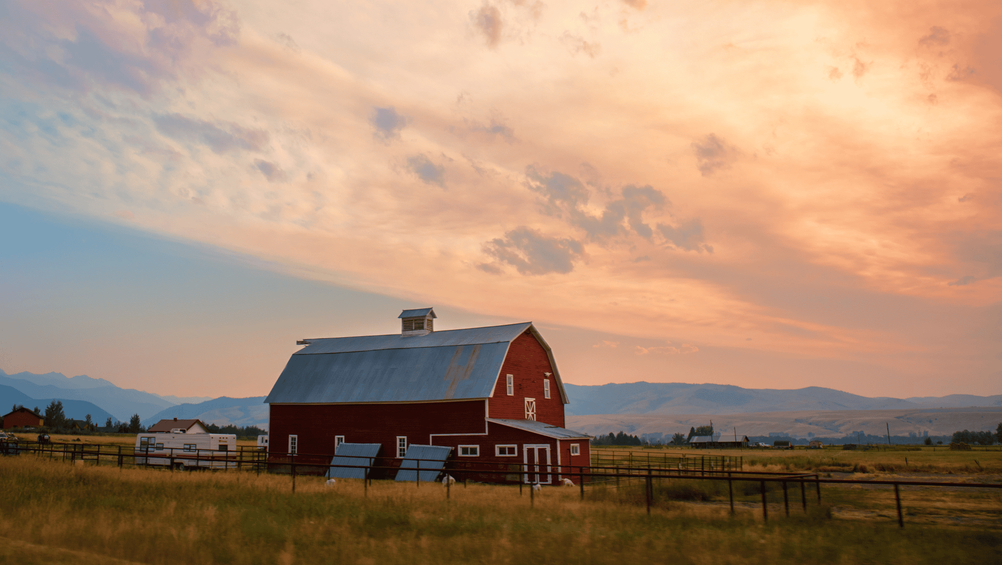 A red barn with a metal roof sits in a grassy field, symbolizing the importance of protecting American farmland, under a colorful, cloudy sky with mountains in the background.