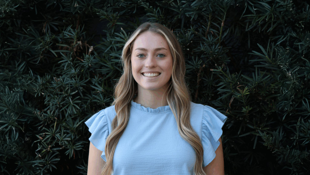 Alecia Dumas, a young woman with long blonde hair, stands smiling in front of dense green foliage, wearing a light blue short-sleeve top.