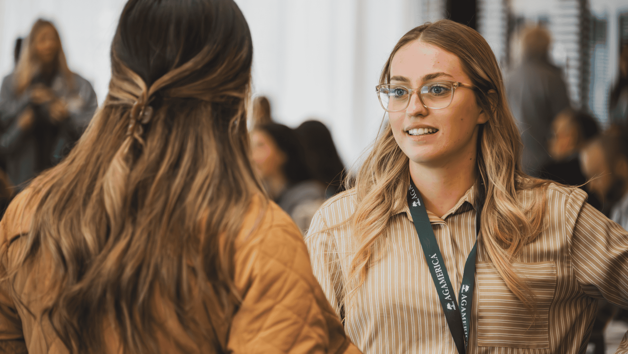 Two women are having a conversation indoors; Alecia Dumas, wearing glasses and a lanyard, faces the camera while the other has her back to it. The scene reflects a professional setting at AgAmerican.
