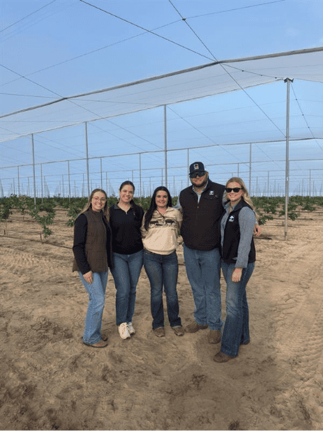 Five people stand together and smile for a photo in a sandy, net-covered agricultural field with young plants.