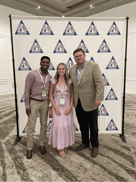 Three people pose in front of a step-and-repeat banner featuring blue and orange triangle logos at an indoor event.