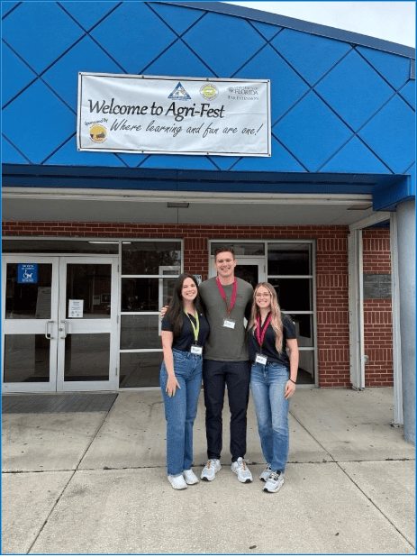 Three people stand in front of a building entrance under a sign that reads "Welcome to Agri-Fest: Where learning and fun are one!.