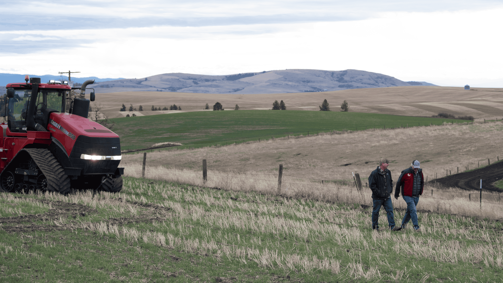 Two people walk through a grassy field near a red tractor, with rolling hills and farmland in the background—an ideal moment to consider farm succession planning and protect your legacy for future generations.